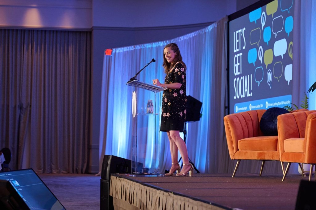 Hannah DeMaio, Vice President of Women Leading Travel, A Skift Brand, in a black floral dress speaks at a podium on a stage with blue lighting. A screen beside her displays "Let's Get Social!" next to colorful speech bubbles.