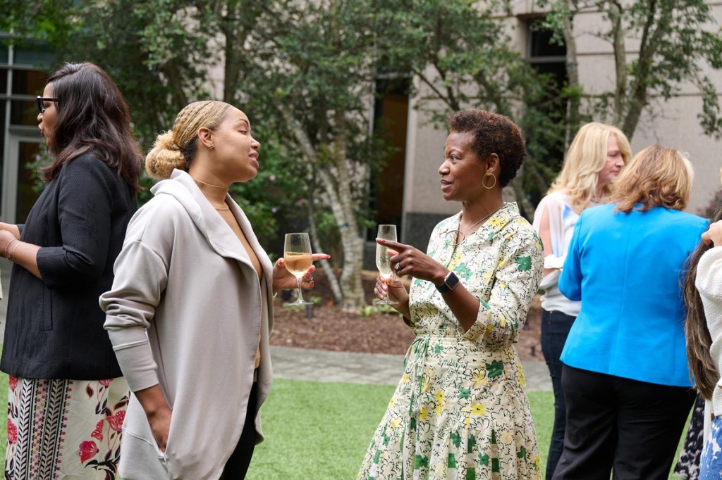 Two women are having a conversation, smiling, and holding glasses of wine at Women Leading Travel event. Three other individuals chat in the background. Trees and green grass create a casual and friendly atmosphere.