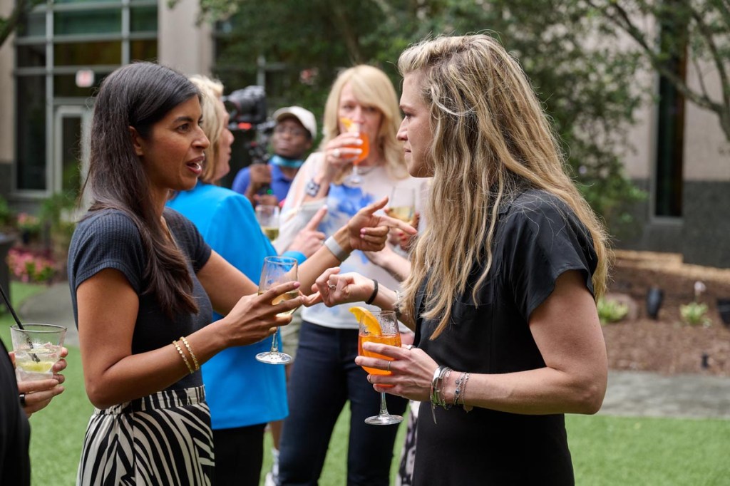 Two women engage in animated conversation, holding cocktails, at an outdoor social gathering. Others in the background enjoy drinks.