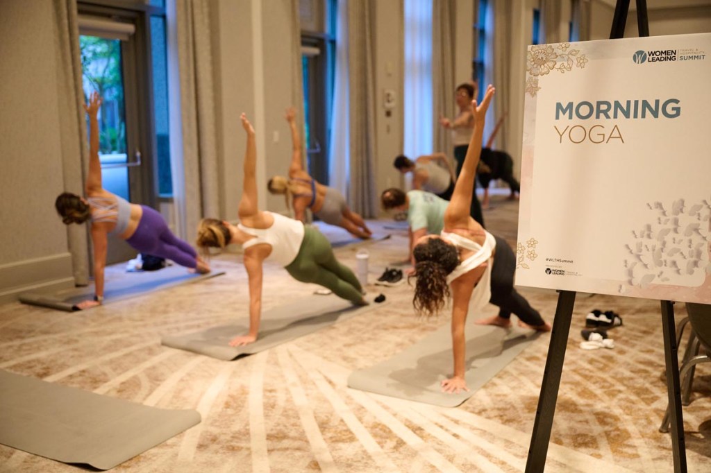 People practice yoga in a serene indoor setting with soft lighting. A sign reads "Morning Yoga." Participants display focus and calmness.