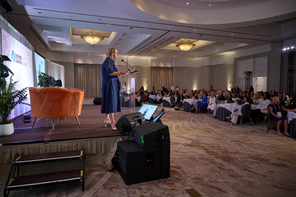 A woman in a blue dress speaks at a podium on a stage for Women Leading Travel event. The room is filled with attentive audience members seated at round tables, creating a formal and engaged atmosphere.