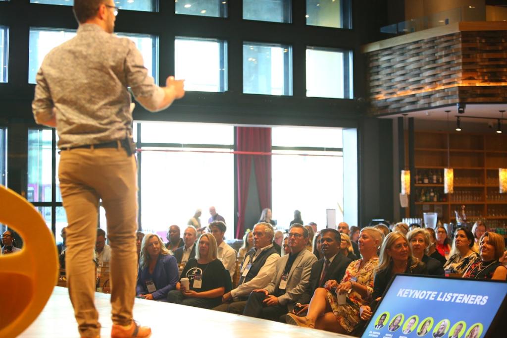 Rear view of a man speaking on a stage in front of a crowd.